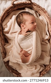 High-angle shot of a tiny newborn infant wrapped in a soft knitted swaddle, lying in a natural wicker basket with neutral-toned linens. The baby is looking to the side with tiny hands and feet visible