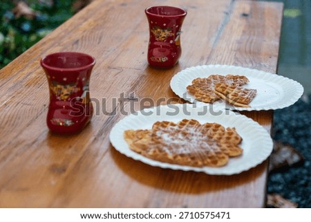 High-angle shot of two red boot-shaped Glühwein mugs and heart-shaped waffles dusted with powdered sugar on a wooden table at a Christmas market. Traditional winter holiday street food and hot drinks 