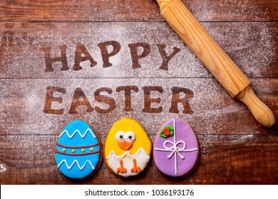 high-angle shot of a wooden table sprinkled with icing sugar or flour where you can read the text happy easter, some cookies decorated as ornamented easter eggs and a funny chick, and a rolling pin