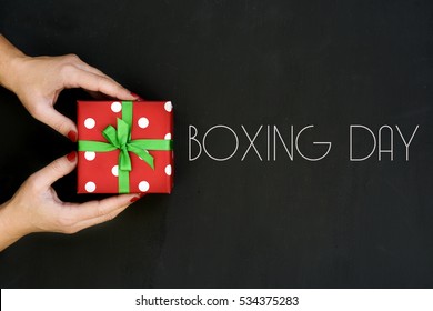 Happy Boxing Day Messages, Wishes, Quotes, And Images 17 high-angle shot of a young caucasian woman with a gift in her hands wrapped in a red paper patterned with white dots and tied with a green ribbon, and text boxing day written on a dark gray surface