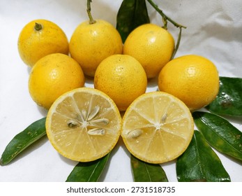 A high-angle, studio-style shot featuring a group of whole, vibrant yellow Meyer lemons and one lemon sliced in half to reveal the juicy pulp and internal seeds. The citrus fruits are arranged naturally with fresh green waxy leaves against a clean white fabric background. This image highlights the organic texture of the citrus peel and the freshness of the fruit, making it ideal for culinary, health, wellness, and food-service marketing materials. The bright yellow color provides a high-contrast, cheerful aesthetic perfect for summer-themed designs or vitamin C related content.