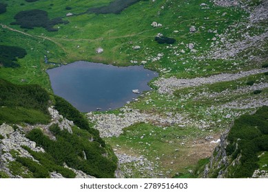 High-angle summer view of a small alpine lake surrounded by grass, rocks, and rugged slopes in the mountains.