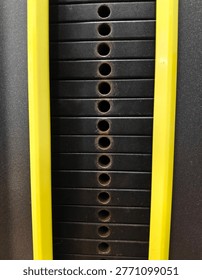 A high-angle, symmetrical close-up of a vertical stack of black metal weight plates in a gym machine, framed by two bright yellow bars. The image emphasizes repetitive patterns and industrial textures.
