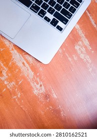A high-angle, top-down shot of a laptop computer placed on a rustic, distressed wooden table. The warm, weathered texture of the wood contrasts with the sleek, modern design of the laptop, conveying themes of remote work, digital lifestyle, creativity, and modern business. Provides ample copy space on the lower area for text or graphic placement.