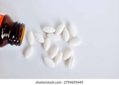 High-angle, top-down view of an open amber glass bottle with white oval medicinal pills poured out onto a bright white background. Flat lay healthcare and pharmaceutical layout.