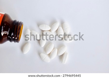High-angle, top-down view of an open amber glass bottle with white oval medicinal pills poured out onto a bright white background. Flat lay healthcare and pharmaceutical layout.