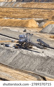 A high-angle, vertical shot capturing a massive industrial spreader machine operating inside the open-pit lignite coal mine in Kostolac, Serbia. The composition illustrates the scale of heavy surface mining equipment surrounded by terraced earth walls, sand mounds, and a long horizontal conveyor belt system. Small mobile cabins and support vehicles are visible next to the crawler-mounted machine, highlighting the immense scale of resource extraction and energy production