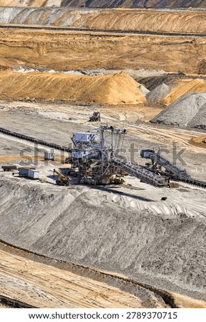 A high-angle, vertical shot capturing a massive industrial spreader machine operating inside the open-pit lignite coal mine in Kostolac, Serbia. The composition illustrates the scale of heavy surface mining equipment surrounded by terraced earth walls, sand mounds, and a long horizontal conveyor belt system. Small mobile cabins and support vehicles are visible next to the crawler-mounted machine, highlighting the immense scale of resource extraction and energy production