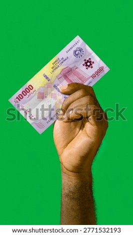 A high-angle, vertical shot features a dark-skinned hand holding a 10,000 Central African CFA franc (XAF) banknote against a solid, vibrant green screen background. The hand, showing the forearm and palm, firmly grips the purple and multicolored bill, which is the 2022 series issued by the Bank of Central African States (BEAC). The note displays intricate security features and the BEAC headquarters. The lighting is bright and even, highlighting the skin texture and the currency's crisp details, ideal for financial or commercial concepts.