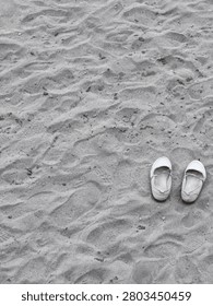 A high-angle, vertical shot featuring a pair of white women's flat shoes carefully placed on an uneven sandy beach surface. The sand background is filled with textured ripples, footprints, and tiny pebbles, creating a serene and relaxed atmosphere. This image perfectly evokes themes of summer vacations, weekend getaways, taking a break, mindfulness, and a carefree beach lifestyle.