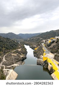 High-angle vertical view overlooking a deep river canyon below a large hydroelectric dam. The image captures the rocky valley, a curved road, and the yellow structures of the dam complex under a dramatic cloudy sky.