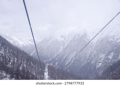 A high-angle view from a cable car looking down at a vast valley and snow-covered mountain ranges, with thick steel ropes cutting through the misty, winter sky.