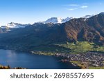 A high-angle view captures a densely settled lakeside town nestled between dark blue water and steep, sun-drenched alpine slopes with snow-capped peaks against a clear sky.