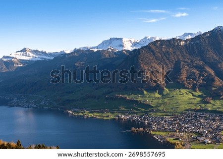 A high-angle view captures a densely settled lakeside town nestled between dark blue water and steep, sun-drenched alpine slopes with snow-capped peaks against a clear sky.