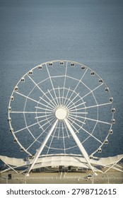 A high-angle view captures the pristine white structure of the Baku Eye Ferris wheel in Azerbaijan against the deep blue, rippling waters of the Caspian Sea. This modern observation wheel stands prominently as an iconic landmark of the city of Baku, offering sweeping panoramic views.