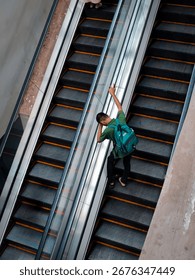 A high-angle view capturing a moment of a young man riding an escalator in a modern building. The man, wearing a green t-shirt and a turquoise backpack, is seen casually leaning against the side. The 
