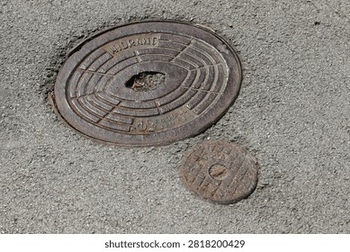 High-angle view of a cast iron hydrant manhole cover and a smaller utility valve cap embedded in grey asphalt pavement.