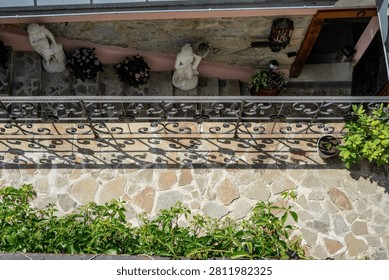 A high-angle view of a charming, multi-level stone terrace featuring ornate wrought-iron railings casting intricate sunlit shadows, accompanied by classical white statues and potted flowers on the lower level.