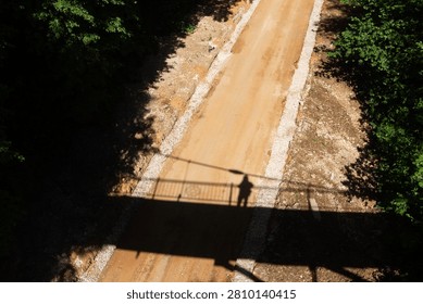 High-angle view of a dusty dirt path beneath a pedestrian bridge casting a dark silhouette of a lone person on the rural road surrounded by lush green summer forest foliage