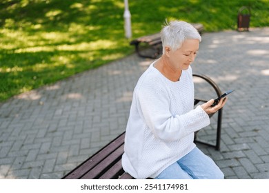 High-angle view of elderly gray-haired woman using smartphone while sitting on bench in public park, enjoying warm weather on sunny summer day. Concept of active aging and happy retirement. - Powered by Shutterstock