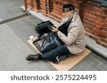 High-angle view of elderly homeless man in hat sitting outside in cold, holding out cup for help in urban street, reflecting issue of homelessness. Concept of a poor unhappy elderly.