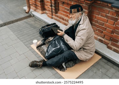 High-angle view of elderly homeless man in hat sitting outside in cold, holding out cup for help in urban street, reflecting issue of homelessness. Concept of a poor unhappy elderly.