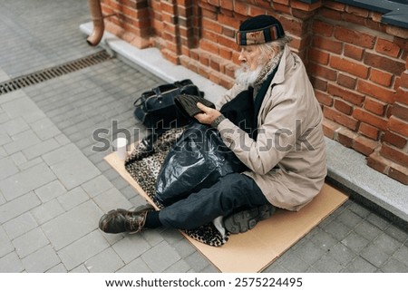 High-angle view of elderly homeless man in hat sitting outside in cold, holding out cup for help in urban street, reflecting issue of homelessness. Concept of a poor unhappy elderly.