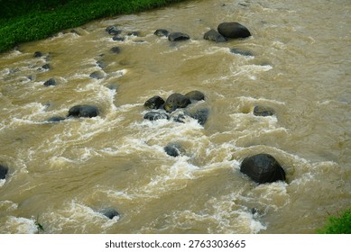 A high-angle view of a fast-flowing, muddy river with turbulent water rushing around large, dark, smooth boulders in a natural outdoor setting.