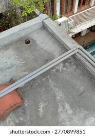A high-angle view of a flat gray concrete rooftop showing parallel metal pipes, a round drain grate, part of a building with white-and-green façade accents, and faint greenery from a tree in the upper left corner.