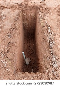 A high-angle view of a freshly dug rectangular grave pit (liang lahad) in reddish-brown soil, with a traditional hoe (cangkul) resting at the bottom, prepared for a burial ceremony.