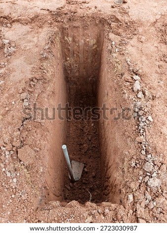 A high-angle view of a freshly dug rectangular grave pit (liang lahad) in reddish-brown soil, with a traditional hoe (cangkul) resting at the bottom, prepared for a burial ceremony.