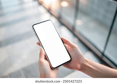 In a high-angle view, a hand holds a mobile smartphone with a blank screen against the blurred background of a modern building walkway outdoors. Business applications, transport, taxi, maps concept - Powered by Shutterstock