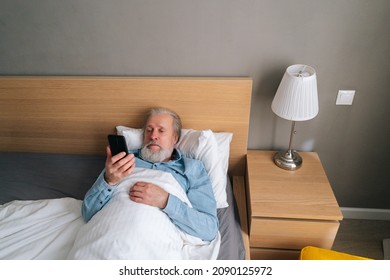 High-angle View Of Handsome Bearded Mature Adult Male Lying On Bed In Hospital And Talking On Mobile Phone By Video Call With Family. Sick Gray-haired Elderly Man Having Meetning In Bed.