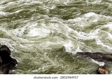 High-angle view looking directly down into the mesmerizing, violent whirlpools and swirling currents of the Jinsha River rapids inside Tiger Leaping Gorge, Yunnan, China. The raging olive-green and foaming white waters churn in a continuous, chaotic cycle against submerged dark stone boulders. Captures the hypnotic, dangerous beauty of extreme river hydrology and raw kinetic energy. An excellent conceptual background for natural forces, chaos, overwhelming power, or turbulent inland waterways.