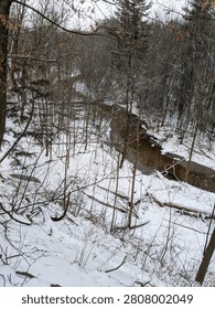 A high-angle view looking down a steep, snow-covered bank at a winding creek flowing through a winter forest. The bare woodland is filled with fallen branches and trees covered in snow.