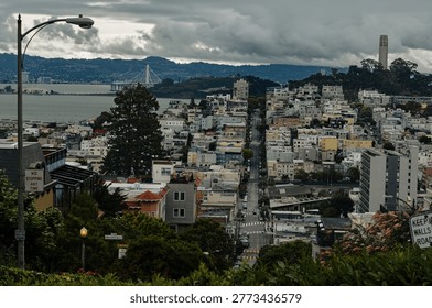 A high-angle view looking east down Lombard Street from the top of the world-famous crooked section. Coit Tower on Telegraph Hill and the San Francisco-Oakland Bay Bridge are visible in the background under a heavy overcast sky.