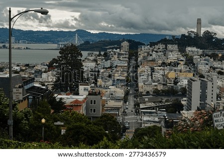 A high-angle view looking east down Lombard Street from the top of the world-famous crooked section. Coit Tower on Telegraph Hill and the San Francisco-Oakland Bay Bridge are visible in the background under a heavy overcast sky.