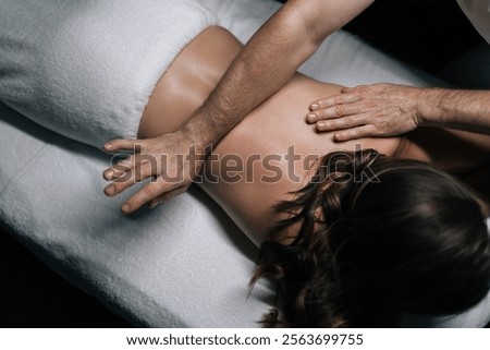 High-angle view of masseur giving back massage to female client lying on massage table in modern spa center with dark interior. Beautiful young woman enjoying relaxing treatment in spa room.