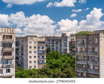 High-angle view of old concrete Soviet-era residential panel buildings in Tbilisi, Georgia. The shot captures the neighborhood surrounded by green trees under a bright blue sky filled with fluffy white clouds, showcasing the contrasting architectural elements and windows.