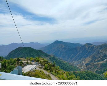 A high-angle view overlooking a deep mountain valley, revealing winding roads carved into the steep slopes framed by green foreground foliage.