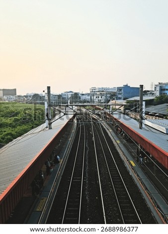 A high-angle view of a railway station with multiple train tracks and platforms, showing commuters waiting for the train during a calm morning in the city. The image captures the perspective lines of 