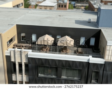 A high-angle view of a rooftop patio featuring wooden picnic tables and two beige canopy tents. The outdoor space is set against a modern building with dark, textured exterior walls.