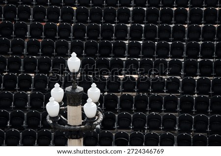 A high-angle view of rows of empty black stadium seats forming a repetitive pattern, with a vintage-style ornate white lamp post in the foreground