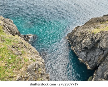 High-angle view of rugged rocky cliffs surrounding clear turquoise water along a dramatic coastal shoreline with textured waves and natural seaside scenery.