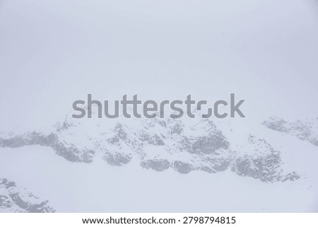 A high-angle view of rugged, rocky mountain slopes covered in snow, shrouded in dense fog and a complete whiteout under overcast winter skies.