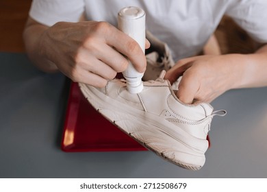 High-angle view of shoemaker using bottle of shoe whitener and painting scuffed surface of dirty white sneaker, illustrating process of cleaning, recoloring, and restoring footwear.