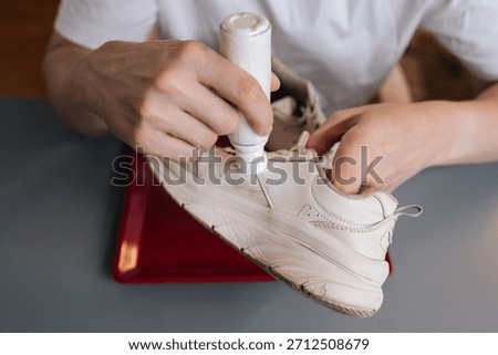 High-angle view of shoemaker using bottle of shoe whitener and painting scuffed surface of dirty white sneaker, illustrating process of cleaning, recoloring, and restoring footwear.
