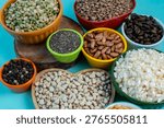 A high-angle view showcasing a diverse collection of raw pantry staples. Various colorful ceramic bowls are filled with green split peas, brown lentils, roasted coffee beans, chia seeds, pinto beans, black peppercorns, white corn, and black-eyed peas, all set against a bright blue surface and a rustic wooden board