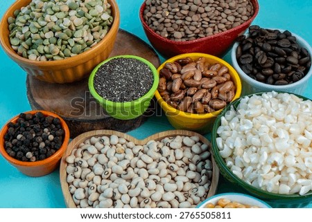 A high-angle view showcasing a diverse collection of raw pantry staples. Various colorful ceramic bowls are filled with green split peas, brown lentils, roasted coffee beans, chia seeds, pinto beans, black peppercorns, white corn, and black-eyed peas, all set against a bright blue surface and a rustic wooden board