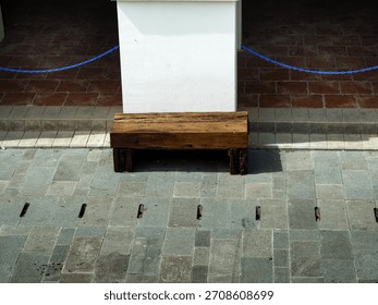 High-angle view of a simple rustic timber bench contrasting with a white column, gray pavement tiles, and a red brick area. Modern urban hardscape design.